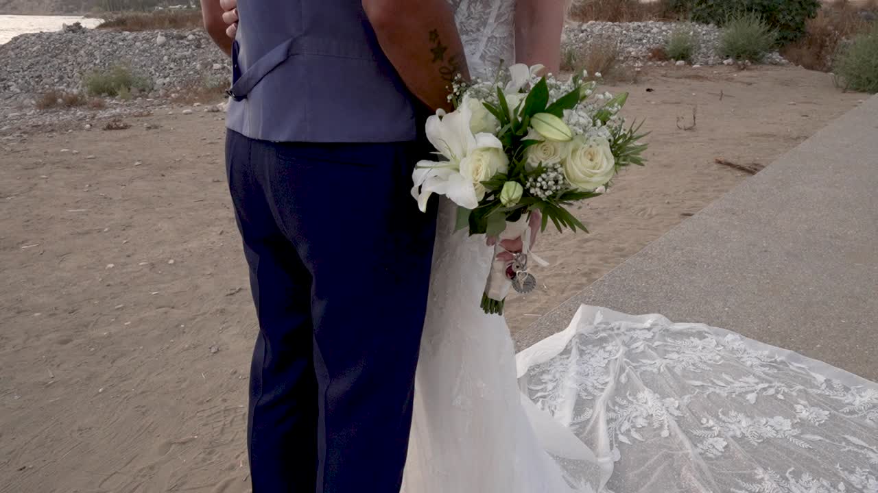 A couple stands together on the beach holding a bouquet of flowers. They are dressed for their wedding with a lace train on the sand. Waves crash in the background as the sun sets