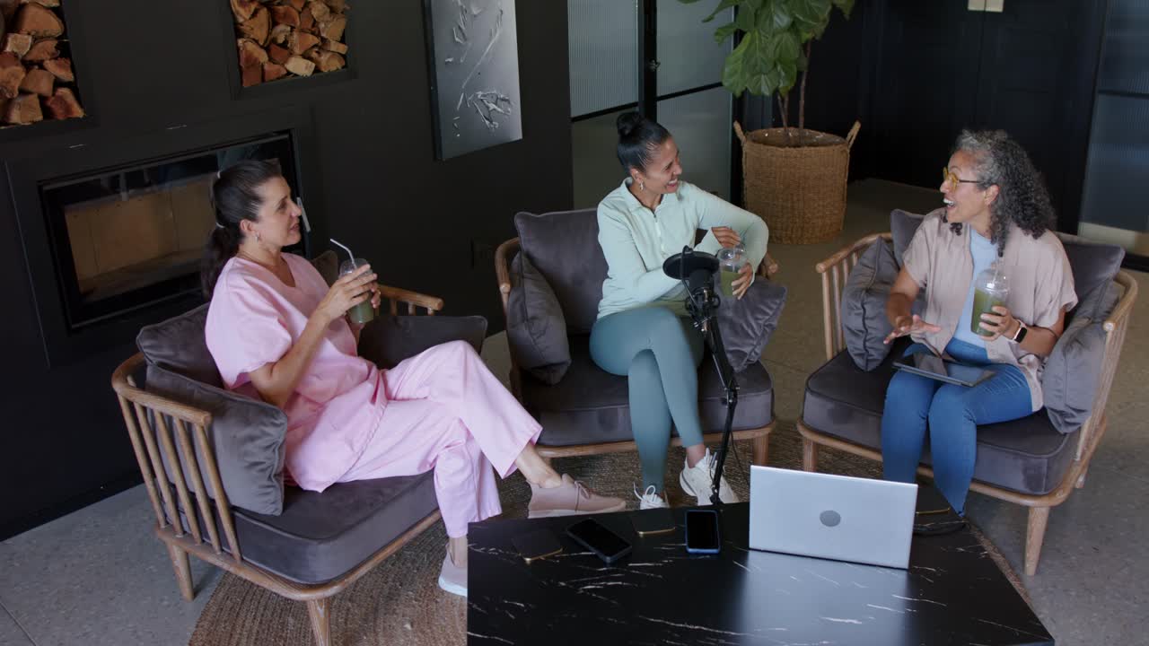 Female friends holding cold drinks at table, center gesturing at mic, sparking right opening laptop