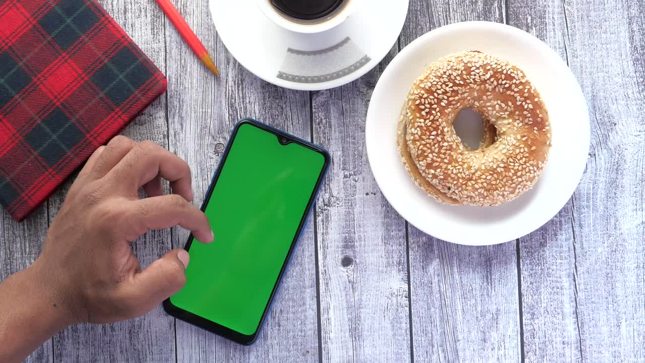 persona usando un teléfono de pantalla verde, café, bagel y cuaderno en una mesa.