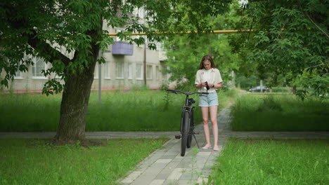 woman holding black gloves while walking up to a parked bicycle on paved path, she put on gloves, wearing smartwatch on right hand, with blurred background of trees and residential building