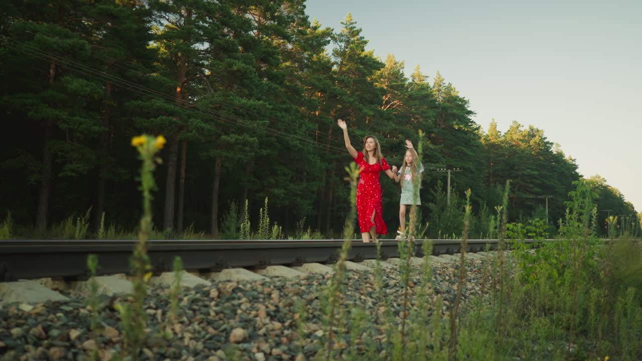 side view of mother in red dress walking hand in hand with daughter waving at someone in distance while balancing on rail track surrounded by forest trees and power lines on bright sunny day