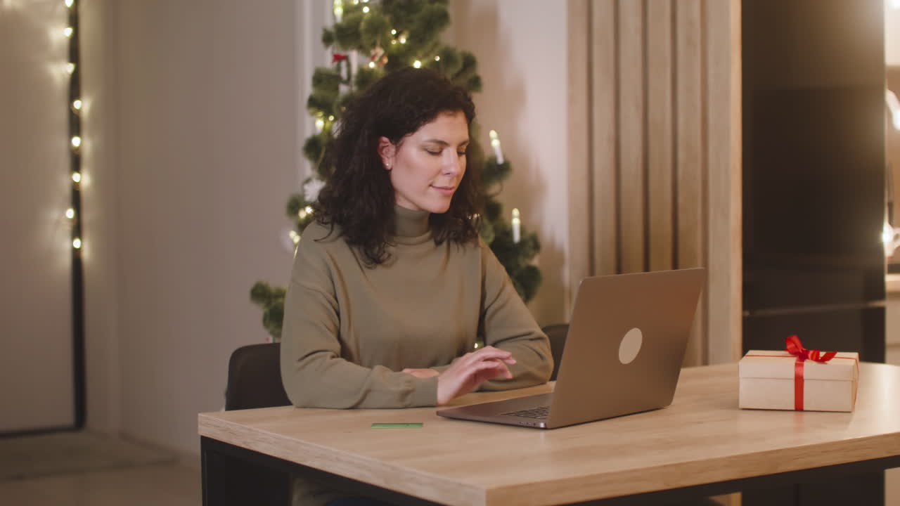 mujer morena comprando en línea con una tarjeta de crédito usando una computadora portátil sentada en una mesa cerca de un regalo en una habitación decorada con un árbol de navidad