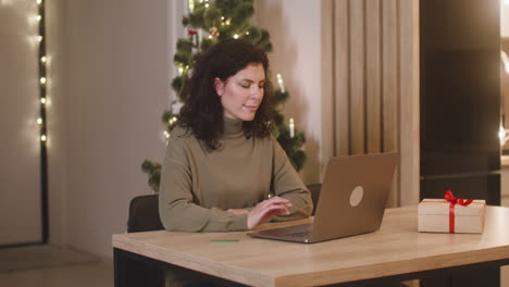 brunette woman buying online with a credit card using a laptop sitting at a table near a present in a room decorated with a christmas tree