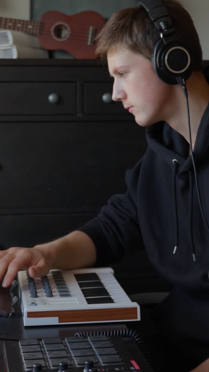 Young man wearing headphones producing music at home, focusing on a MIDI keyboard, composing a new song in his private studio, developing creative sound, using laptop touchpad, vertical panning