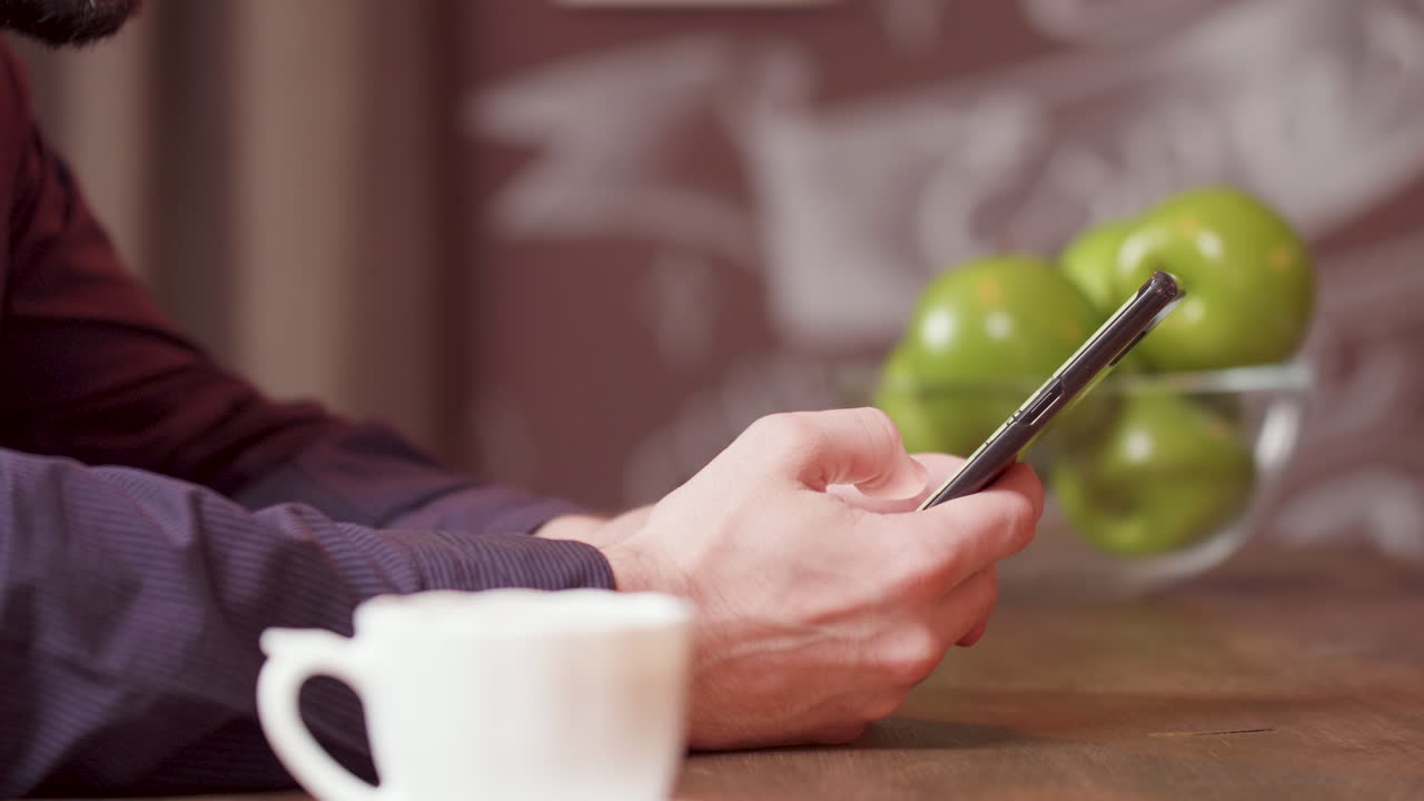 Close up shot of a man using a smartphone at a wooden table with coffee and apples in background