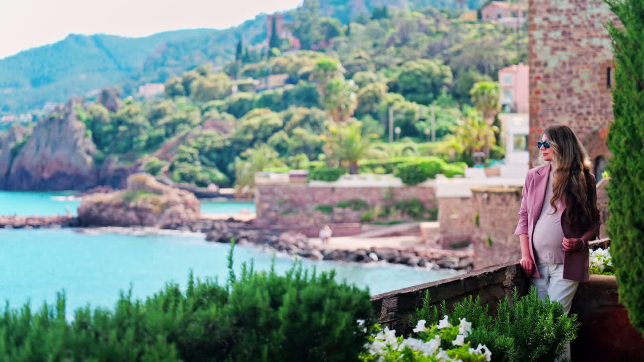 Pregnant woman with long hair, wearing sunglasses standing with the Chateau de la Napoule Castle in Mandelieu-La Napoule, France in the background