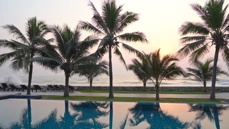 pan over a resort swimming pool lined with palm trees, ocean waves roll in as the sunsets in the background