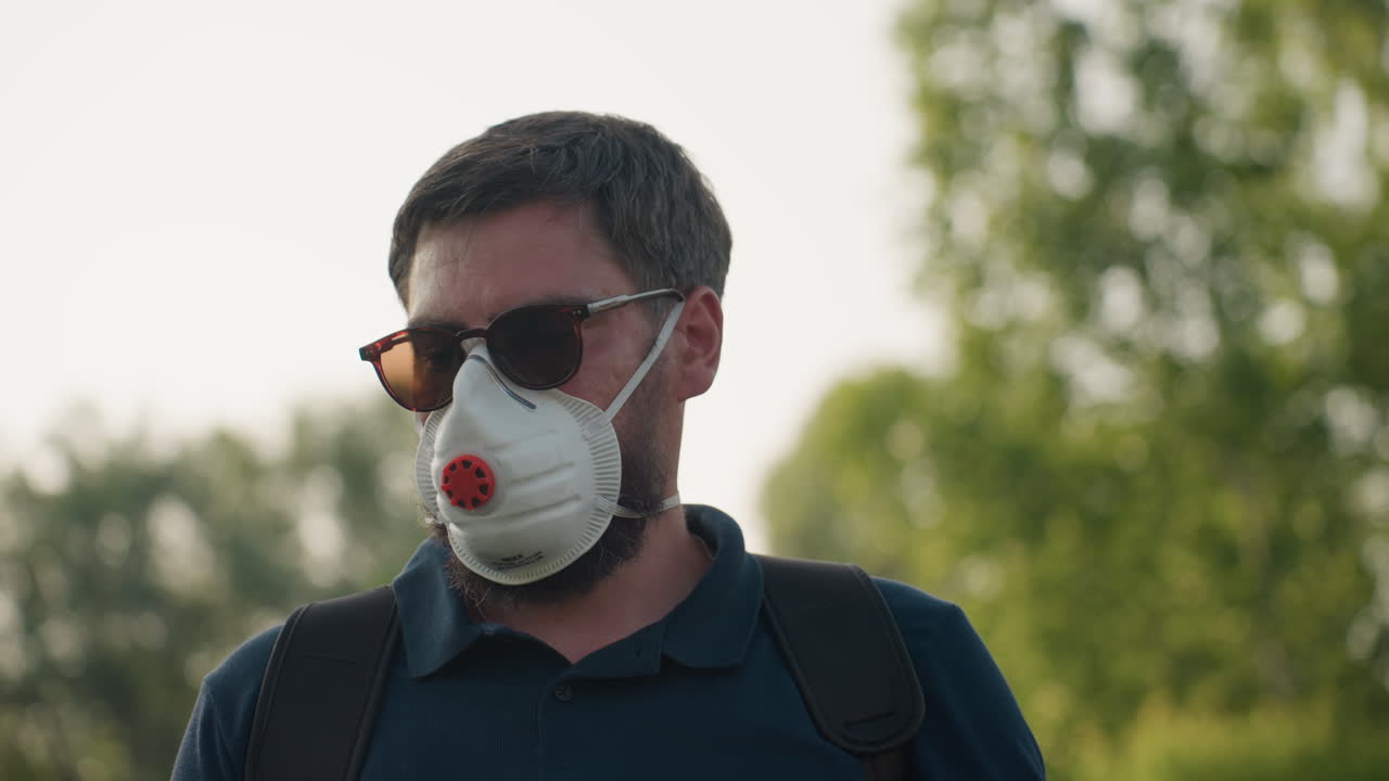 Close up of man wearing protective face mask and sunglasses adjusting backpack strap in outdoor setting with trees in background, under natural light