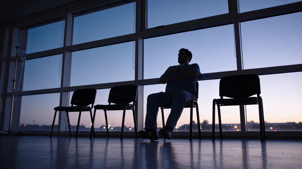 Uomo seduto in divisa medica in pausa, spostando lo sguardo e incrociando le braccia alla finestra dell'ospedale, con un'asta per flebo.