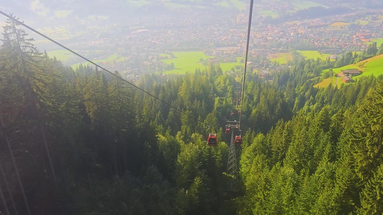 Kitzbühel, Austria, corsa in funivia sopra una verde valle alpina, con linee di impianti di risalita, tetti di villaggi, foresta montana e paesaggio turistico estivo soleggiato nella regione del Tirolo.