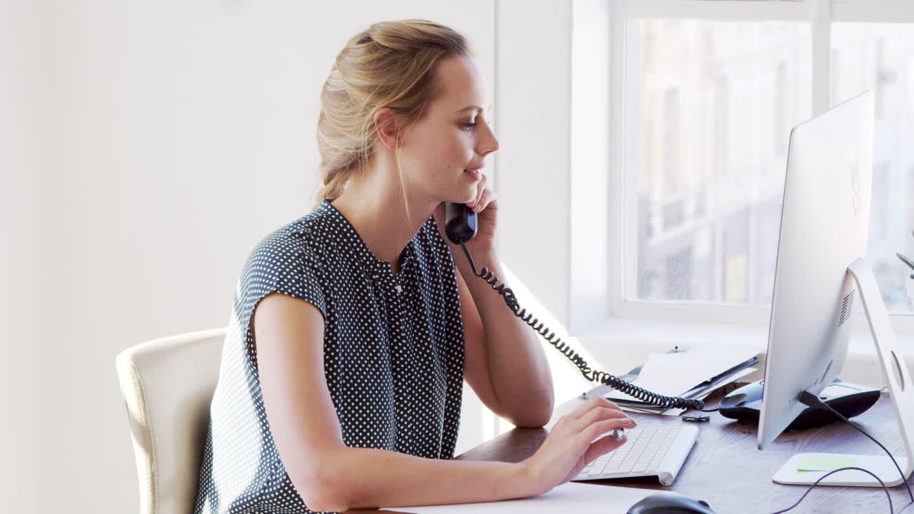Ung hvit kvinne bruker telefon og datamaskin på et kontor