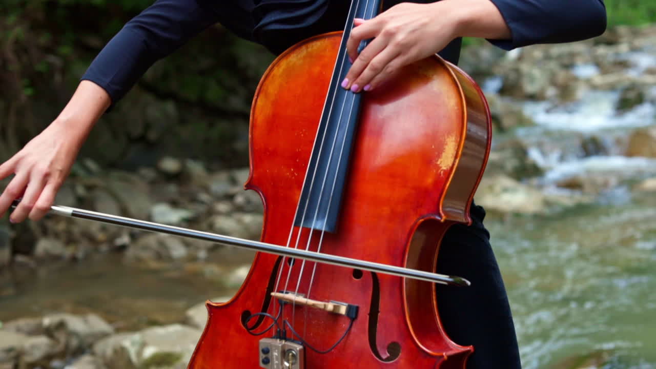 Woman plays the cello outdoors. Female musician playing the instrument on natural river background in summer. Musical equipment in womans' hands.