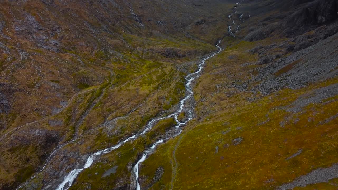 Aerial drone footage flying over and above a small narrow river or river rapids with white water that is flowing in grassy brown rocky scottish hills and mountains during a cloudy day time.