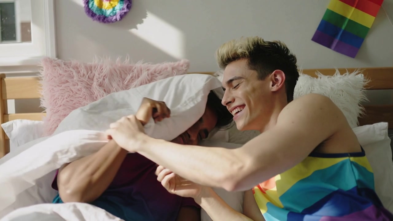 Uomo dalla pelle chiara che afferra un cuscino, due uomini che giocano/lottano sul letto in camera da letto con una canotta arcobaleno.