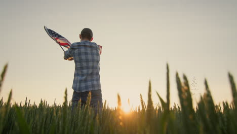 american farmer waving the us flag against the backdrop of a field of wheat where the sun rises. low angle shot