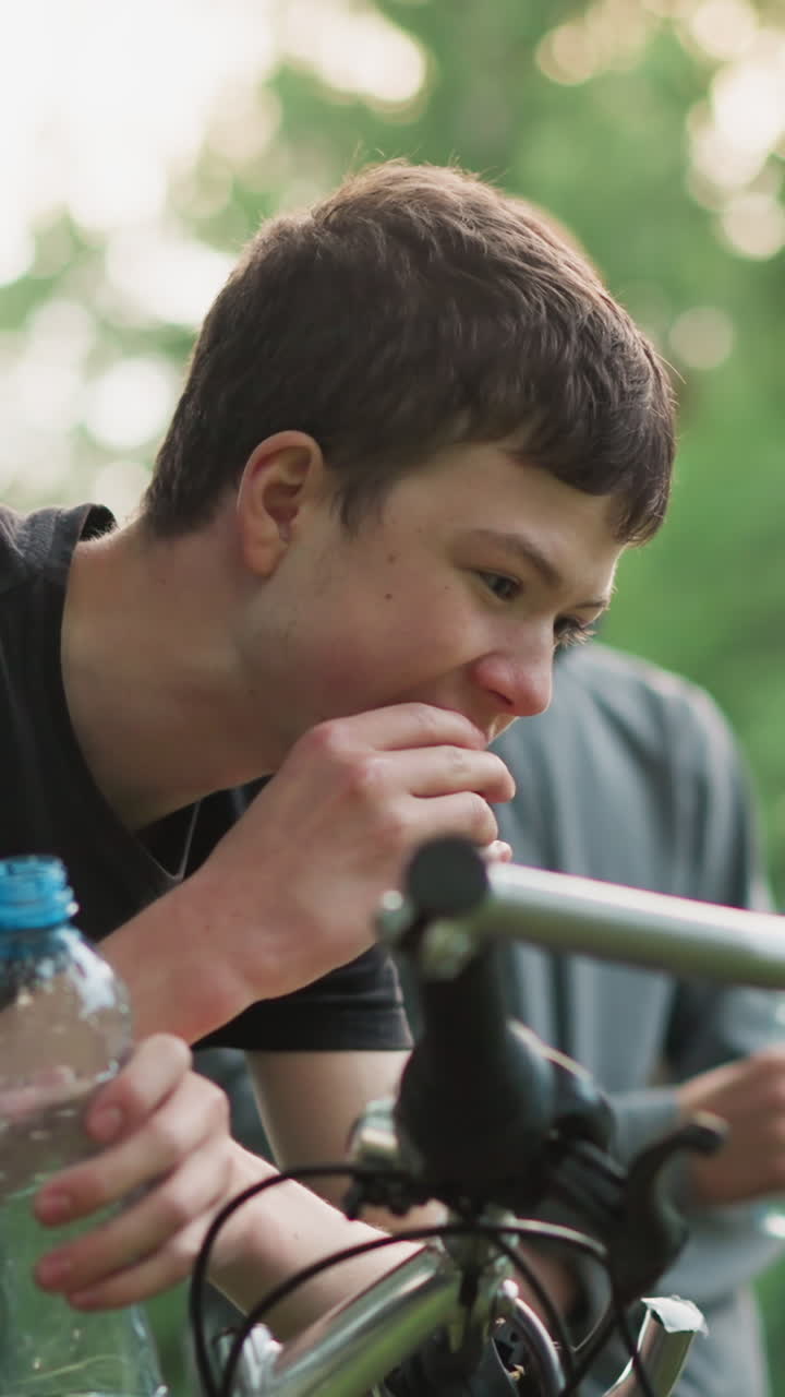 어린 소년은 맛있는 간식과 물 병을 즐기면서 자전거 손잡이에 기대어, 다른 사람이 물을 마시면서, 배경에는 흐릿한 초록색이 있습니다.
