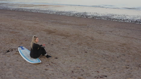 drone shot of woman wearing wetsuit sitting on surfboard and looking out to sea