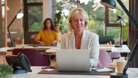 mature businesswoman working on laptop at desk in office pausing to look out of window