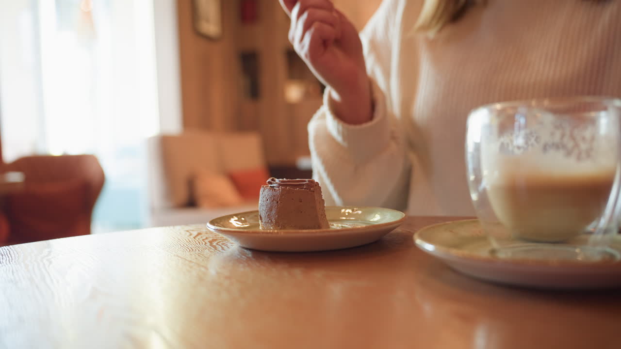 close up of customer in cafeteria gently slices rich chocolate dessert with fork beside half filled latte glass, cozy interior with soft blur background