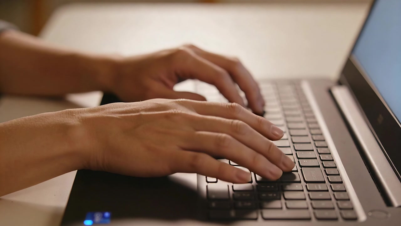 Starting from rest, person's hands typing across laptop keyboard at desk for work, blue LED glowing