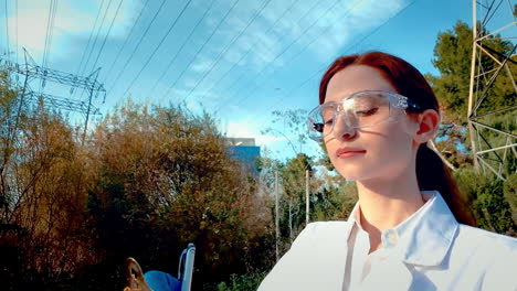 A-young-woman-scientist-at-a-creek,-wearing-protective-eyewear-and-a-lab-coat,-taking-notes-on-a-clipboard