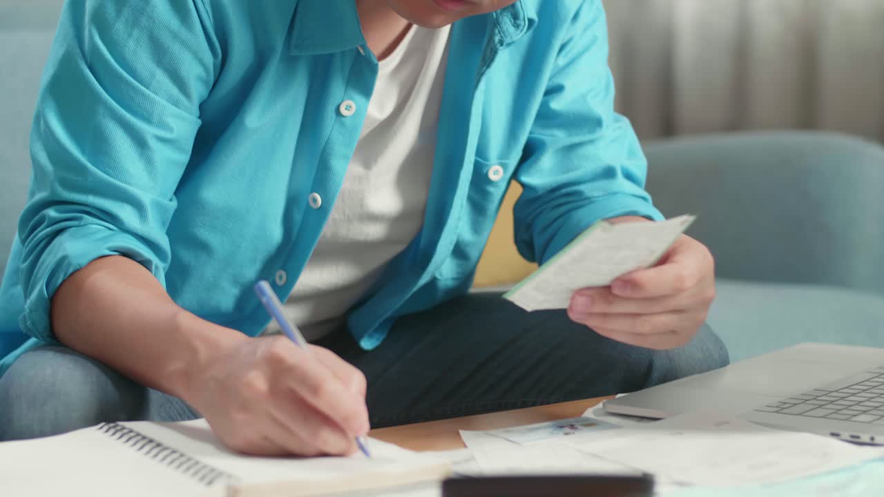 Primo piano di un uomo asiatico con un portatile che tiene in mano e guarda la bolletta mentre registra le spese sul quaderno.