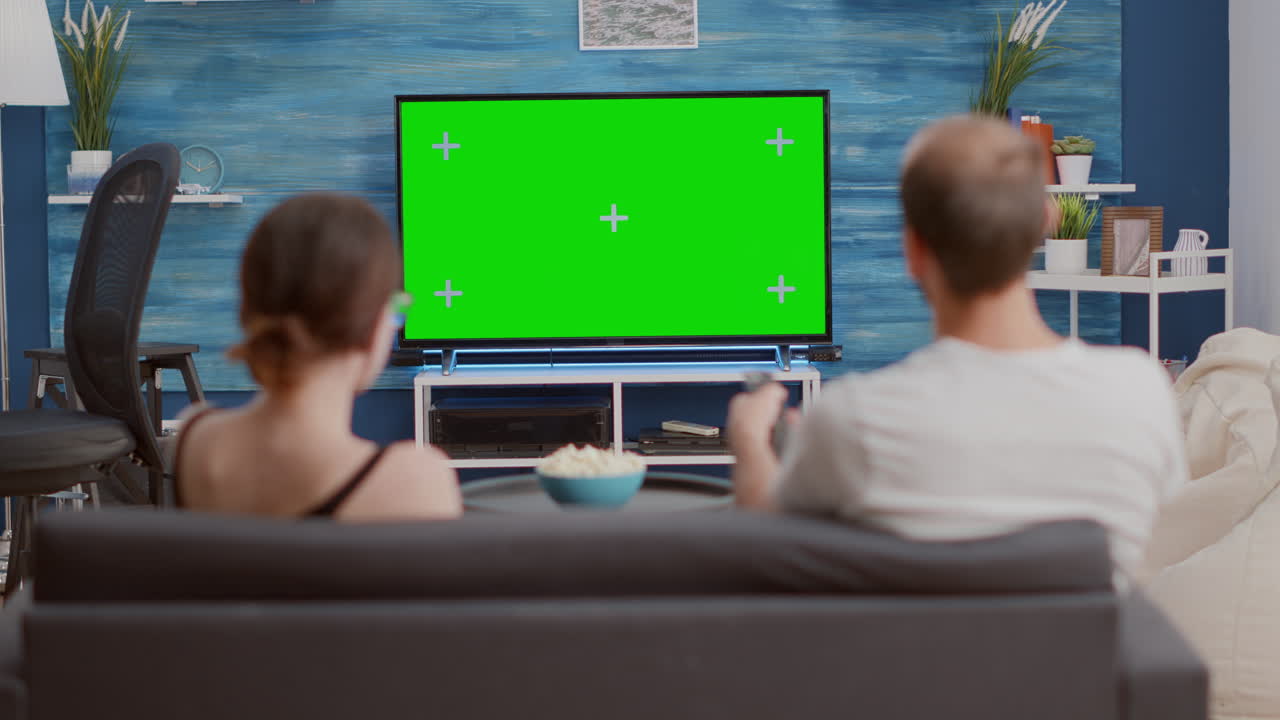 Couple sitting on couch watching television on green screen flat tv talking about favourite show and zapping
