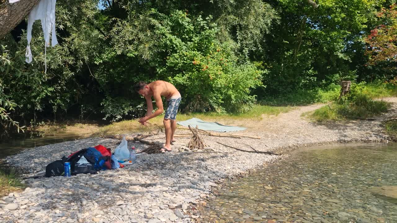 Attraktiv mand i badetøj forbereder træ til en grillfest ved Aare-flodbredden i Münsingen, Bern, Schweiz, og nyder en perfekt sommerdag udendørs.