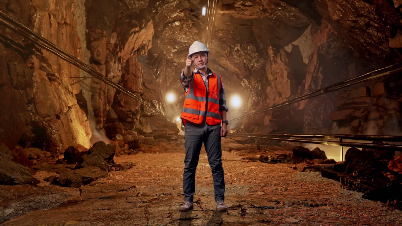 Full Body Of Asian Male Engineer With Safety Helmet Smiling And Touching His Chest Then Pointing At You While Standing In Underground Mine Tunnel