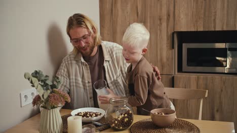 A-little-albino-boy-with-white-hair-and-blue-eyes-pours-milk-into-a-plate-for-his-father,-a-blond-man-with-a-beard-and-glasses-in-a-white-checkered-shirt-during-their-breakfast-in-a-modern-kitchen