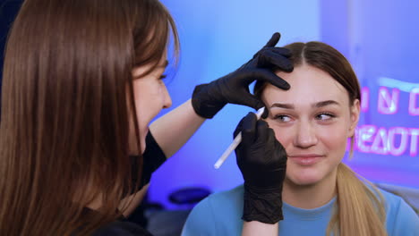 brunette long-haired beautician marks the eyebrow lines for the young beautiful girl. preparation for eyebrow make up. close up.