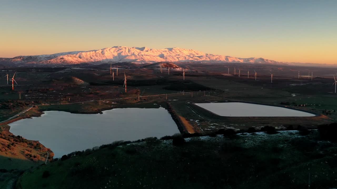 Sunrise or Sunset over Snow-capped Mountains with Wind Turbines and a Reservoir
