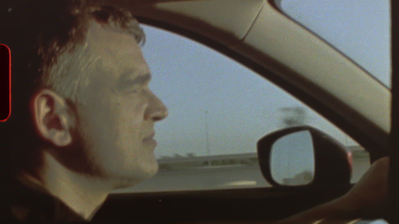 Filmed on super 8 film Middle aged driver navigating highway lane, vintage 8mm film aesthetic capturing side profile through car window with nostalgic grainy texture and cinematic movement