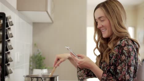 Smiling-young-woman-uses-smart-phone-in-kitchen-while-cooking-on-stove,-dolly-shot