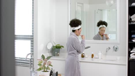 Biracial-woman-in-bathrobe-brushing-teeth-and-using-smartphone-in-bathroom,-slow-motion