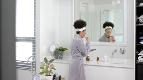 mujer biracial en bata de baño cepillándose los dientes y usando un teléfono inteligente en el baño, cámara lenta