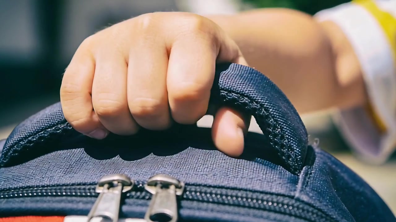 Small child's hand in yellow sleeve gripping padded blue backpack handle outside, preparing to lift