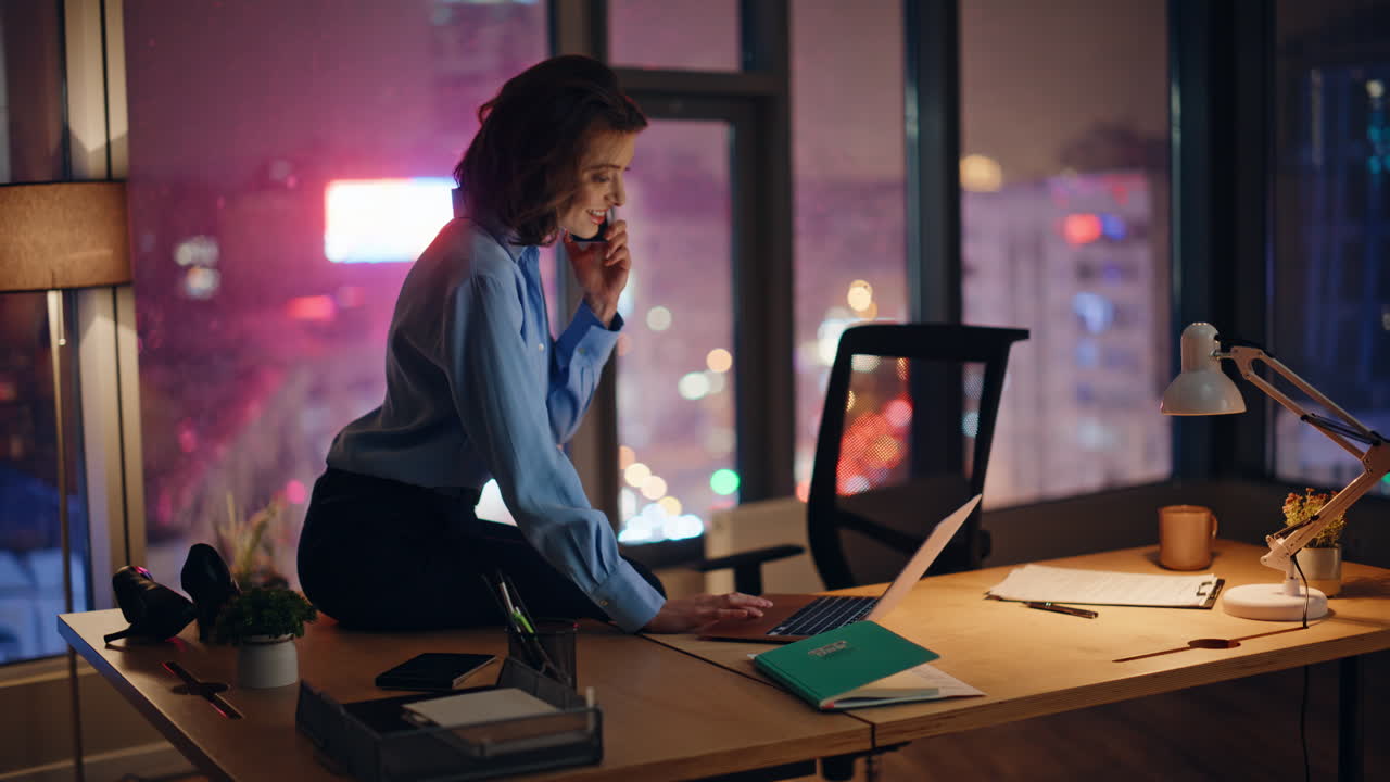 Happy workaholic talking mobile phone sitting at dark work desk looking laptop