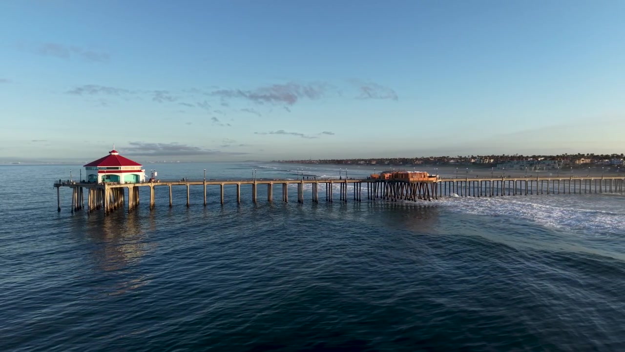 Il drone scivola lentamente lungo il lato dell'iconico molo di Huntington Beach all'alba. Le onde si infrangono contro i suoi possenti piloni mentre si erge maestoso contro l'Oceano Pacifico