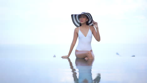 Foto-Clave-Alta-De-Una-Hermosa-Joven-Asiática-Con-Un-Gran-Sombrero-Sentado-En-El-Borde-De-La-Piscina-Infinita