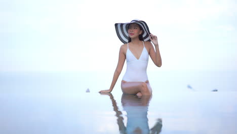 high key shot of beautiful young asian woman with big hat sitting in infinity pool edge