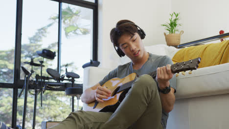 asian boy wearing headphones playing guitar in living room at home