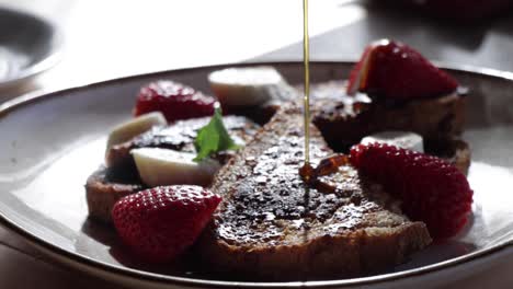 Organic-honey-being-poured-on-whole-bread-French-toasts,-including-sliced-bananas-and-strawberry-fruit,-in-a-low-light-table