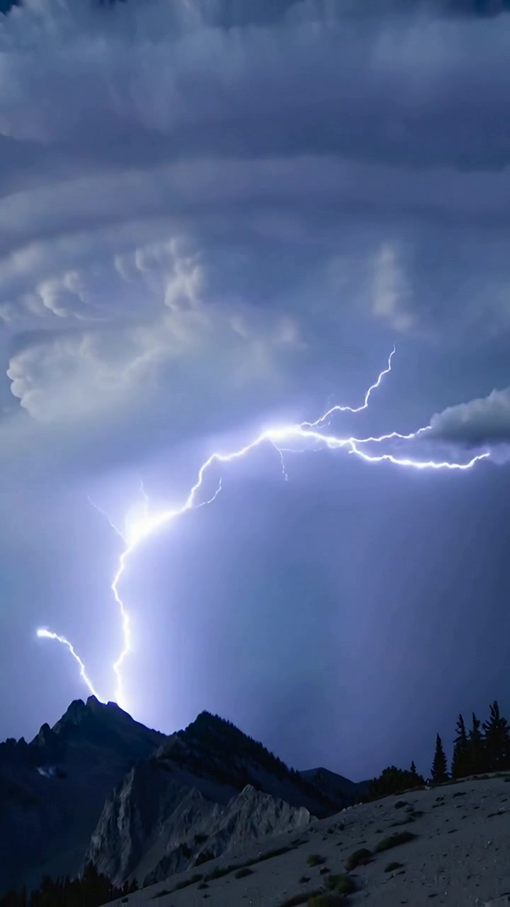 Video verticale: Nuvola temporalesca lampeggiante che rilascia fulmini, illuminando cime frastagliate e una cresta di conifere