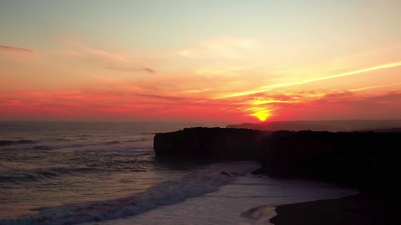 A red sky during sunset over the rocky cliffs while strong waves crash on the sandy beach near London Bridge rocks on Victoria's Great Ocean Road