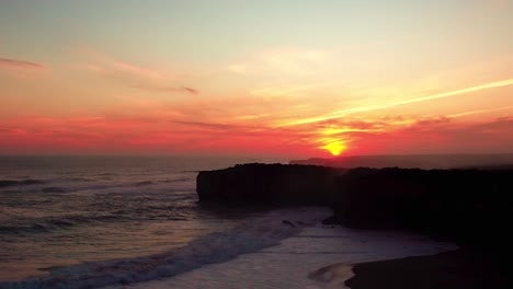 Un-Cielo-Rojo-Durante-La-Puesta-De-Sol-Sobre-Los-Acantilados-Rocosos-Mientras-Fuertes-Olas-Rompen-En-La-Playa-De-Arena-Cerca-De-Las-Rocas-Del-Puente-De-Londres-En-La-Gran-Carretera-Oceánica-De-Victoria