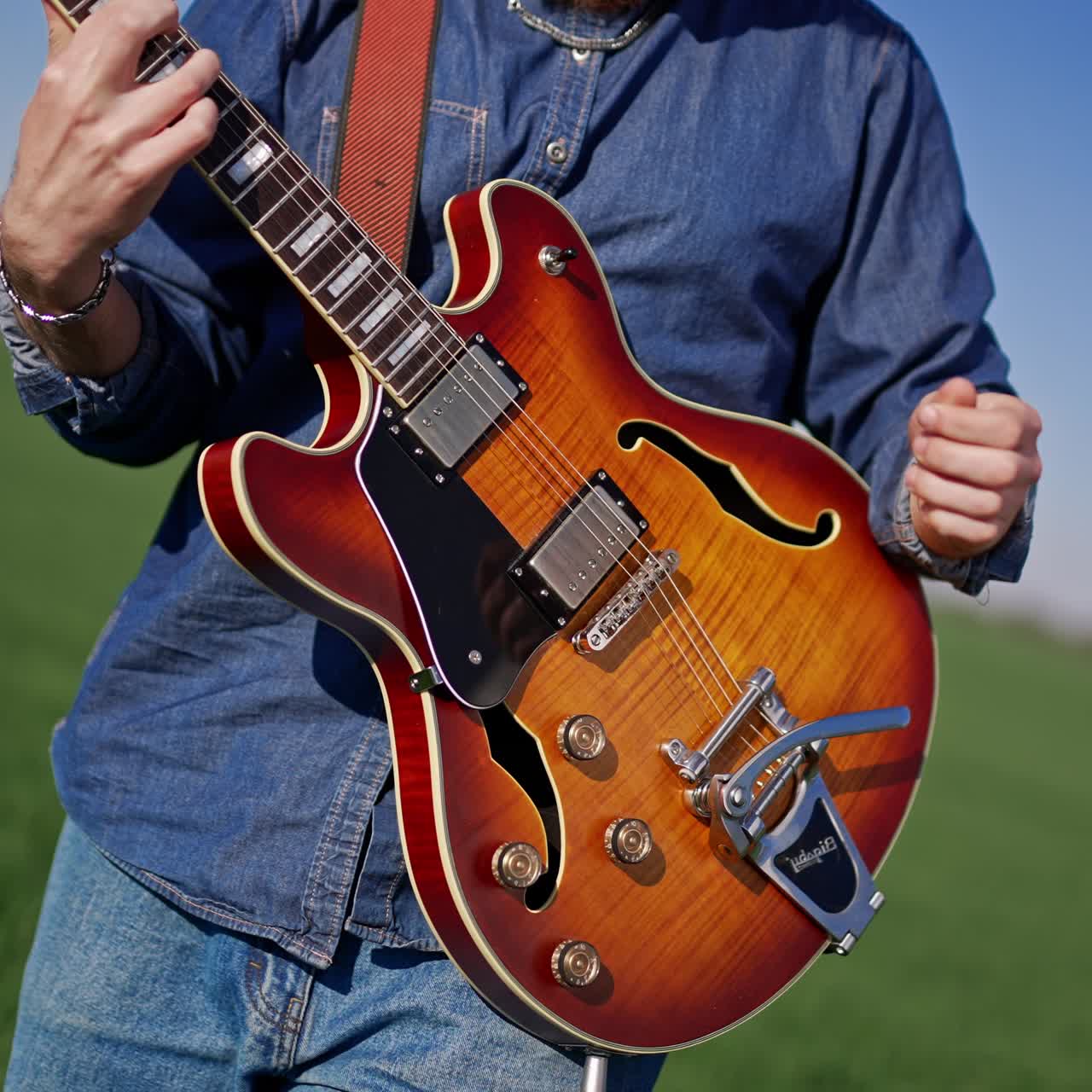 Stylish young bearded man playing guitar in the beautiful farmland. Musician playing drums at backdrop in blur