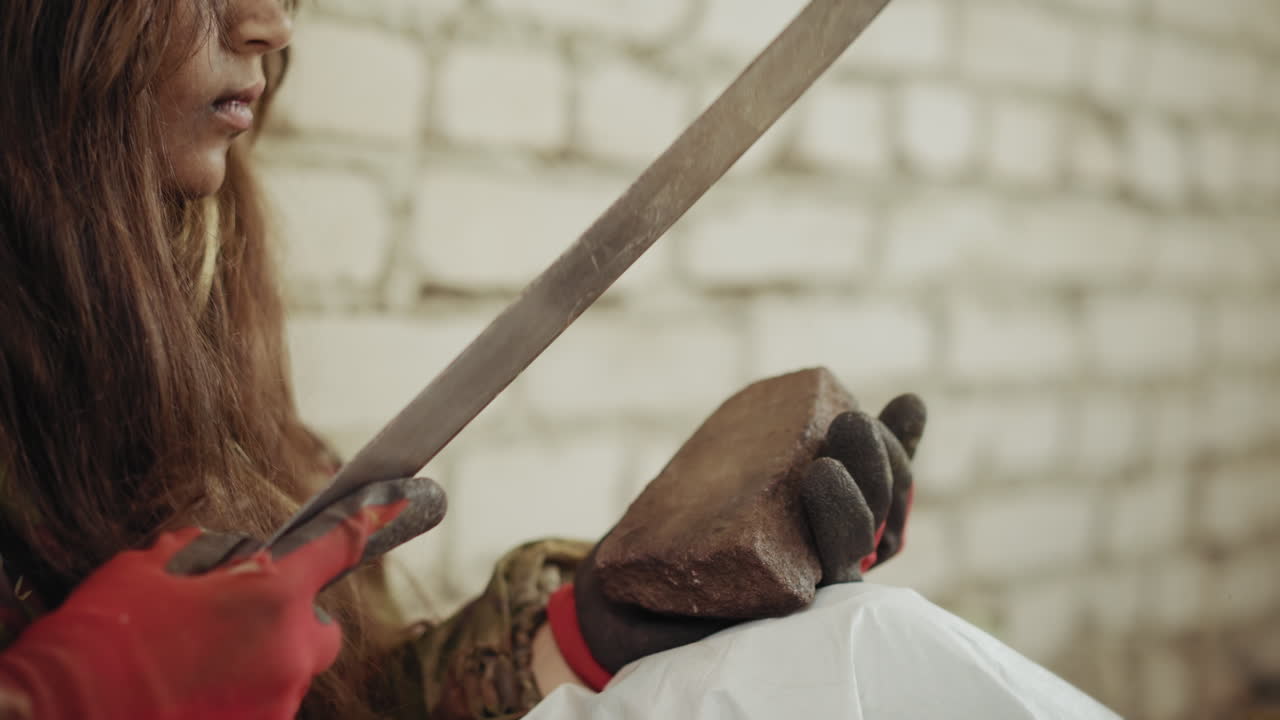 Close up of orphan sharpening cutlass blade on stone with gloved hand, face lowered in concentration as sharp metal edge presses firmly against rough block