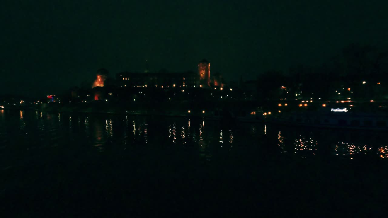 Lo skyline di una città illuminata è visibile di notte. L'acqua riflette queste luci, aggiungendo una profondità interessante. Le persone si godono la vista lungo la riva del fiume.