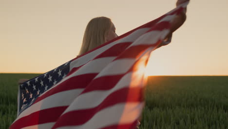 handheld shot: a woman with an american flag on her shoulders watches the sun go down over a field of wheat.